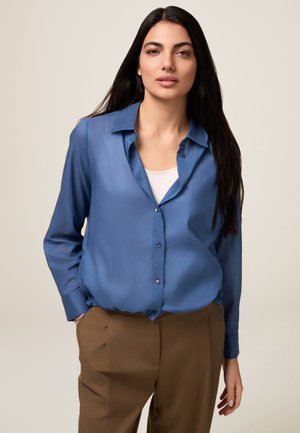 Camicia azzurra chiaro con una texture liscia, maniche lunghe e colletto morbido, abbinata a pantaloni marroni.