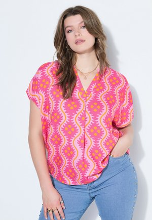 Kobieta o falowanych brązowych włosach, ubrana w żywą różową wzorzystą bluzkę i jasnoniebieskie dżinsy z ćwiekami, pozująca z jedną ręką w kieszeni na tle białej ściany.