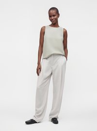 Femme debout avec une main dans la poche, portant un haut gris clair sans manches, un pantalon blanc à jambes larges et des chaussures noires sur fond blanc.