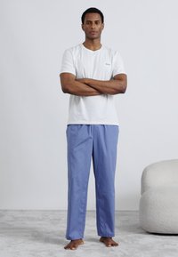 T-shirt bianca a maniche corte con un logo discreto, abbinata a pantaloni azzurri, in piedi a piedi nudi su una superficie morbida e strutturata.