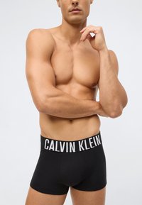 Svarta boxerkalsonger med en bred midjeband som har "CALVIN KLEIN" i vit skrift. Slät tyg, nära passform, inga ytterligare mönster.