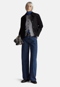 Černá semišová bunda, šedý cardigan na knoflíky, modrý rolák, široké džíny a černé mokasíny. Drží károvaný šátek.