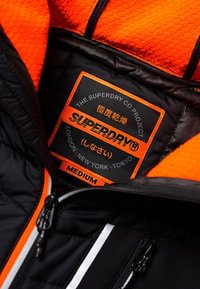 Innenraum einer schwarzen und orangefarbenen Jacke mit hexagonalem Muster. Enthält ein Etikett mit dem Markennamen "Superdry", der Größe "Medium" und Städtenamen.