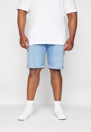 Mężczyzna w jasnoniebieskich zwiniętych dżinsowych szortach, białych skarpetkach do kostek, białych sneakersach i prostym białym t-shircie z krótkim rękawem stoi na białym tle.