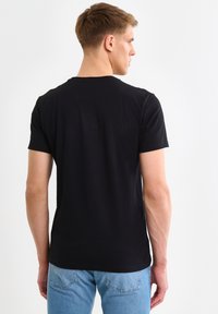 Giovane uomo di spalle che indossa una t-shirt nera a maniche corte semplice e jeans azzurri chiari su sfondo bianco uniforme.