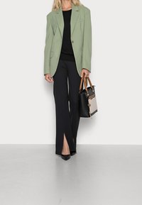 Hellgrüner übergroßer Blazer, schwarzes Oberteil, hoch taillierte schwarze Hose mit seitlichen Schlitzen, schwarze spitze Schuhe und eine schwarze Handtasche mit beigen Akzenten.