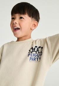 Sweat-shirt couleur crème avec une finition texturée, présentant un graphique de trois pingouins et le texte "PENGUIN PARTY" en bleu marine.