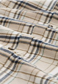 Beige geruite stof met bruine, blauwe en groene lijnen. Zachte textuur met zichtbare stiksel en een knoopdetail. Ideaal voor kleding of accessoires.