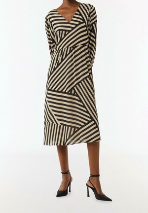 Femme portant une robe midi rayée noire et beige à manches longues, avec un décolleté en V et des chaussures noires à talons hauts avec bride à la cheville.