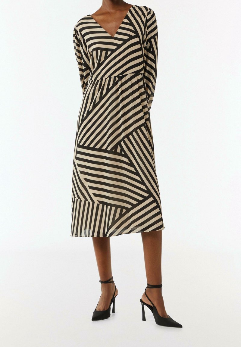 Femme portant une robe midi rayée noire et beige à manches longues, avec un décolleté en V et des chaussures noires à talons hauts avec bride à la cheville.