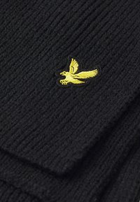 Črna pletena kapa z teksturiranim rebrastim vzorcem. Ima rumeni vezeni logotip ptice na sprednji strani. Mehak, fleksibilen material.