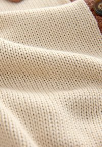 Tissu tricoté couleur crème avec des coutures serrées et régulières et des plis subtils créant une surface douce et texturée.