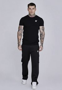 Musta lyhythihainen t-paita, jossa rintalogo, yhdistettynä mustiin lukuisiin taskuihin varustettuihin cargo-housuihin ja valkoisiin lenkkareihin.