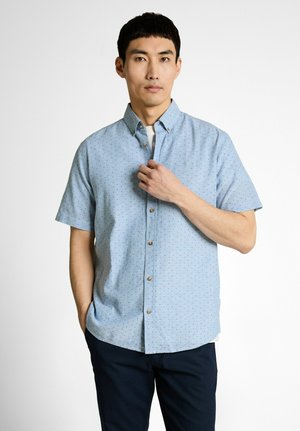 Man wearing light blue short-sleeve button-up shirt with small print, dark pants, standing against plain white background.