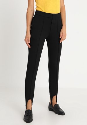 Zwarte broek met een aansluitend ontwerp, met een hoge taille, zijsplitten aan de zoom en een gladde textuur. Gecombineerd met loafers en een geel top.