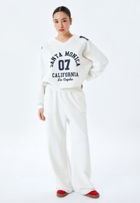 Felpa bianca con testo navy "SANTA MONICA 07 CALIFORNIA Los Angeles," abbinata a pantaloni della tuta bianchi e scarpe rosse, vestibilità rilassata, tessuto morbido.