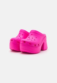 Roze platformne klompe, izdelane iz lahkega materiala, z ventilacijskimi luknjami, ukrivljeno zasnovo in teksturirano podplat, ki zagotavlja oprijem.