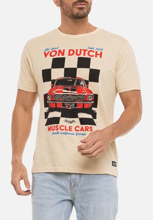 Homme portant un t-shirt beige avec un muscle car rouge et un drapeau à damier noir, associé à un jean bleu clair.