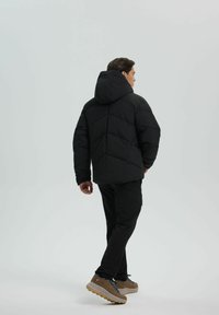 Schwarze Puffjacke mit Kapuze, gestepptem Muster und gerader Silhouette. Kombiniert mit schwarzen Hosen und braunen Sportschuhen.