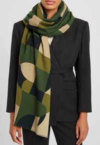 Sciarpa verde e beige con motivo, realizzata in tessuto morbido, con forme geometriche, bordi frangiati, appoggiata casualmente intorno al collo.