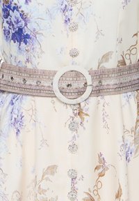 Robe à motif floral avec des tons lavande et beige, décorée de boutons ronds scintillants et d'une ceinture texturée avec une grande boucle circulaire.