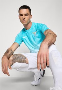 Muž s tetováním, oblečený v modrém sportovním dresu, bílých šortkách, ponožkách a kopačkách, se sklání a dívá se do kamery na jednobarevném pozadí.