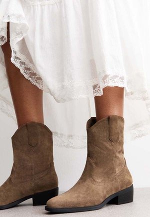 Bruine suède enkelboots met een gestapeld hak, spitse neus en decoratieve stiksel. Draagt met een witte rok met kant hem.