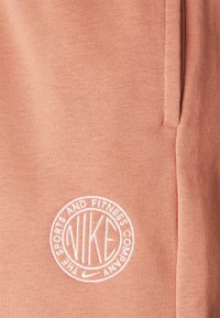 Nike sweatpants i mjuk korallfärgad tyg med en slät textur. Har en vit broderad logotyp i en cirkulär design nära fickan.