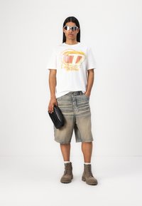 Hvit grafisk t-skjorte med fargerik trykk, falmet blå denimshorts og brune snørestøvler. Tilbehør inkluderer en svart clutch og sølvsolbriller.
