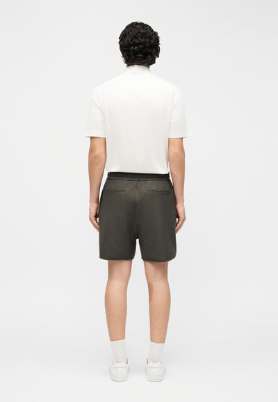 Άνδρας με σγουρά σκουρόχρωμα μαλλιά, που στέκεται πλάτη, φοράει λευκό κοντομάνικο πουκάμισο, σκούρα σορτς, λευκές κάλτσες και λευκά αθλητικά παπούτσια, σε λευκό φόντο.