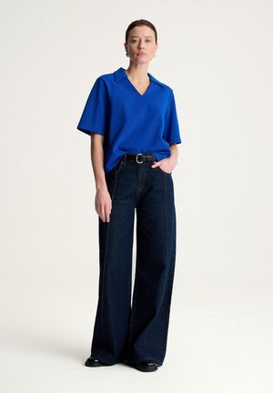 Kobieta stojąca z ręką w kieszeni, ubrana w niebieski top z krótkim rękawem i dekoltem w serek oraz szerokie, ciemne dżinsy z czarnym paskiem i butami.