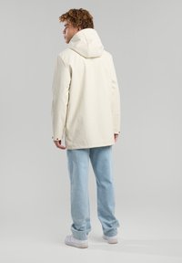 Chaqueta impermeable color beige claro con capucha, mangas largas y ventilación trasera. Llevada sobre unos vaqueros de mezclilla azul claro y zapatillas blancas.