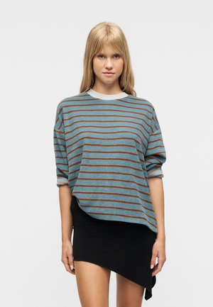 Mladá žena s blond vlasmi nosí modrý a hnedý pruhovaný sveter a asymetrickú čiernu mini sukňu, stojaca na bielom pozadí.