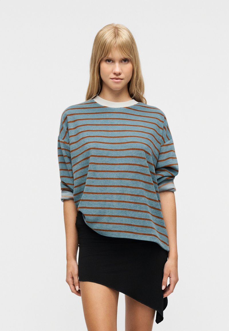 Νεαρή γυναίκα με ξανθά μαλλιά φοράει πουλόβερ με μπλε και καφέ ρίγες και ασύμμετρη μαύρη μίνι φούστα, στέκεται μπροστά σε λευκό φόντο.