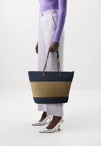 Sac en toile navy matelassé avec un fond en paille tissée et des anses en chaîne dorée, arborant un logo, tenu à côté de pantalons lavande pâle.