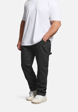 Mand iført en almindelig hvid T-shirt, sorte cargobukser med lommer og hvide sneakers, stående med den ene hånd i lommen.