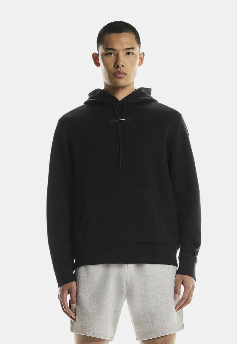 Zwarte katoenen hoodie met een voorzak, capuchon met trekkoord en geborduurd labeldetail. Model draagt lichtgrijze shorts.
