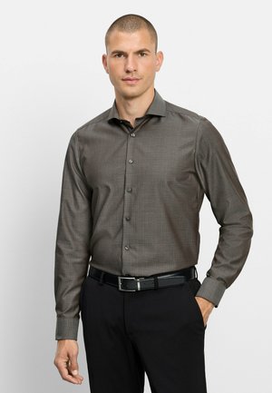 Mann mit kurzen Haaren, der ein braunes kariertes Langarmhemd trägt, das in schwarze Hosen mit schwarzem Gürtel gesteckt ist, vor weißem Hintergrund.