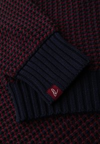 Neulepaita, jossa on teksturoitu kuosi tummansinisessä ja burgundin punaisessa, ja joustavat hihat. Punainen etiketti logolla, joka on näkyvissä hihassa.