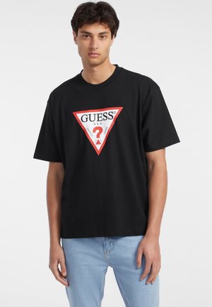Musta puuvillainen t-paita lyhyillä hihoilla, jossa on suuri punainen kolmionmuotoinen logo, jossa lukee "GUESS U.S.A." ja valkoinen kysymysmerkki sisällä.