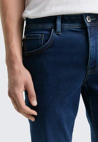 Vaqueros de denim azul oscuro con un diseño clásico de cinco bolsillos, costuras en contraste y cierre de botón visible. Textura suave.
