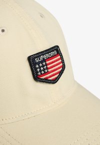Light beige cap featuring a navy badge with "SUPERDRY" and a red, white, and blue embroidered American flag design.