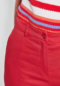 Rote Baumwollhosen mit glatter Textur, ausgestattet mit einem Vorderknopf und seitlichen Taschen, kombiniert mit einem mehrfarbigen gestreiften Strickoberteil.