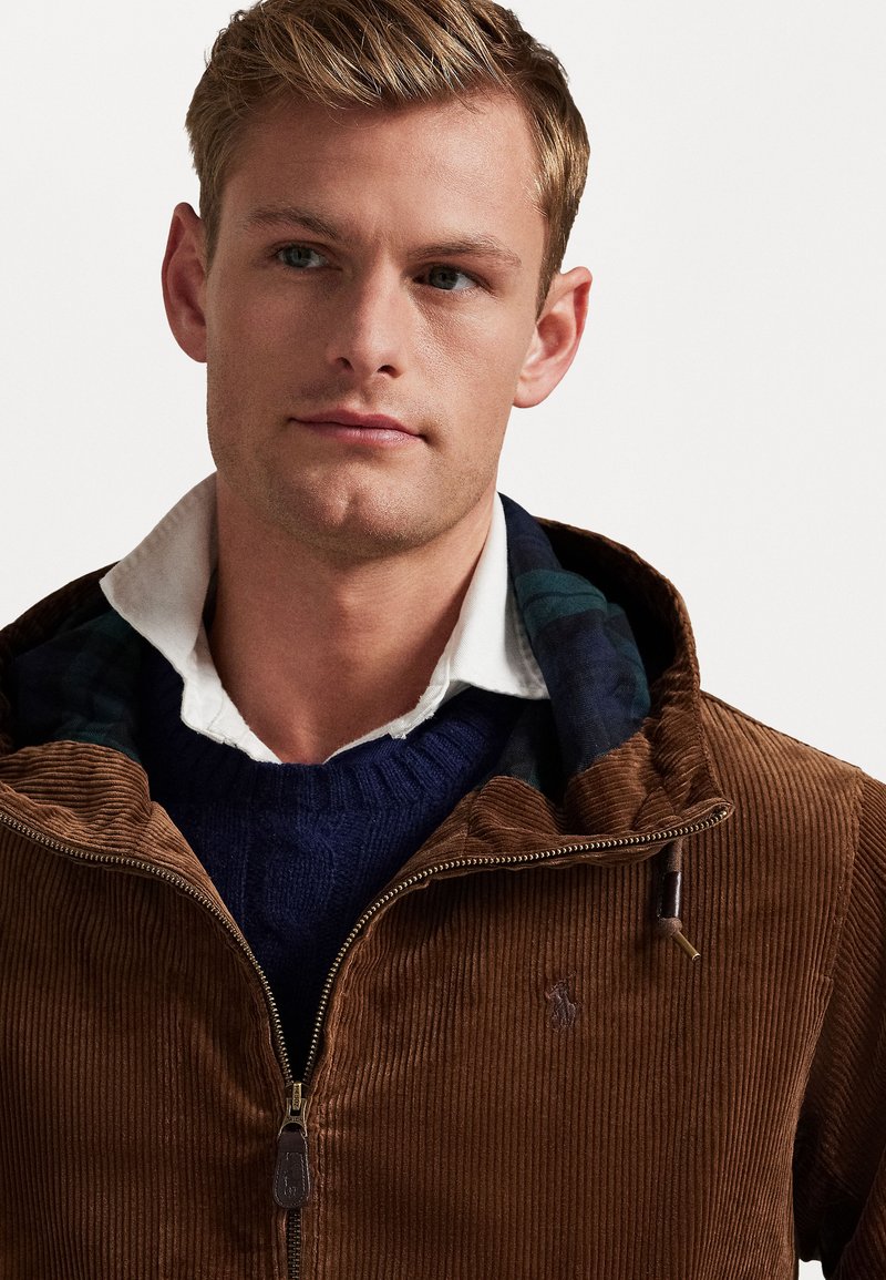 Giacca in corduroy marrone con zip frontale, abbinata a un maglione blu a coste e una camicia bianca con colletto sotto. Dettagli includono una fodera a quadri.