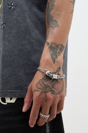 Hopeaketju, jossa on pääkalloaihe ja kiinnityskohta, on tatuoidussa kädessä. Sormuksessa on vastaava pääkallo sormus nimettömässä.