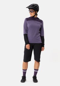 Maglietta da ciclismo a maniche lunghe viola con maniche nere, abbinata a pantaloni corti neri e calze a righe viola. Indossata con un casco nero e occhiali da sole.