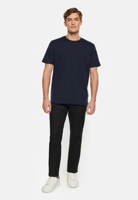 Dunkelblaue Baumwoll-T-Shirt mit Rundhalsausschnitt, kurzen Ärmeln und einem kleinen Logodetail auf der Brust, kombiniert mit schwarzen Slim-Fit-Hosen und weißen Sneakers.