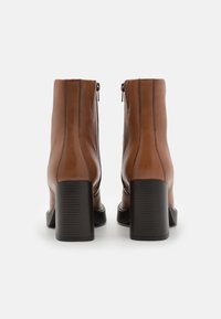 Botas de tobillo de cuero marrón con una textura suave, punta redonda y tacón cuadrado. Presentan un cierre con cremallera en la parte trasera.
