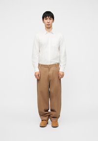 Λευκό πουκάμισο με κουμπιά, καφέ παντελόνι με φαρδιά μπατζάκια και ανοιχτό καφέ παπούτσια. Διαθέτει τσέπη στο στήθος και άνετη εφαρμογή.