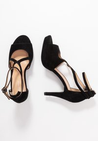 Czarne zamszowe sandały na wysokim obcasie z odkrytymi palcami, z krzyżującymi się paskami i cienkim obcasem, leżące na białym tle.